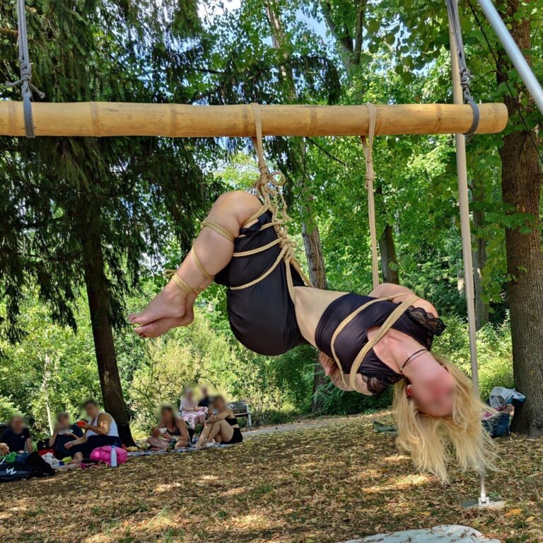 Hängefesselung am Bambus zwischen Bäumen im Park, das blonde Haar hängt herab, im Hintergrund Picknickgäste
