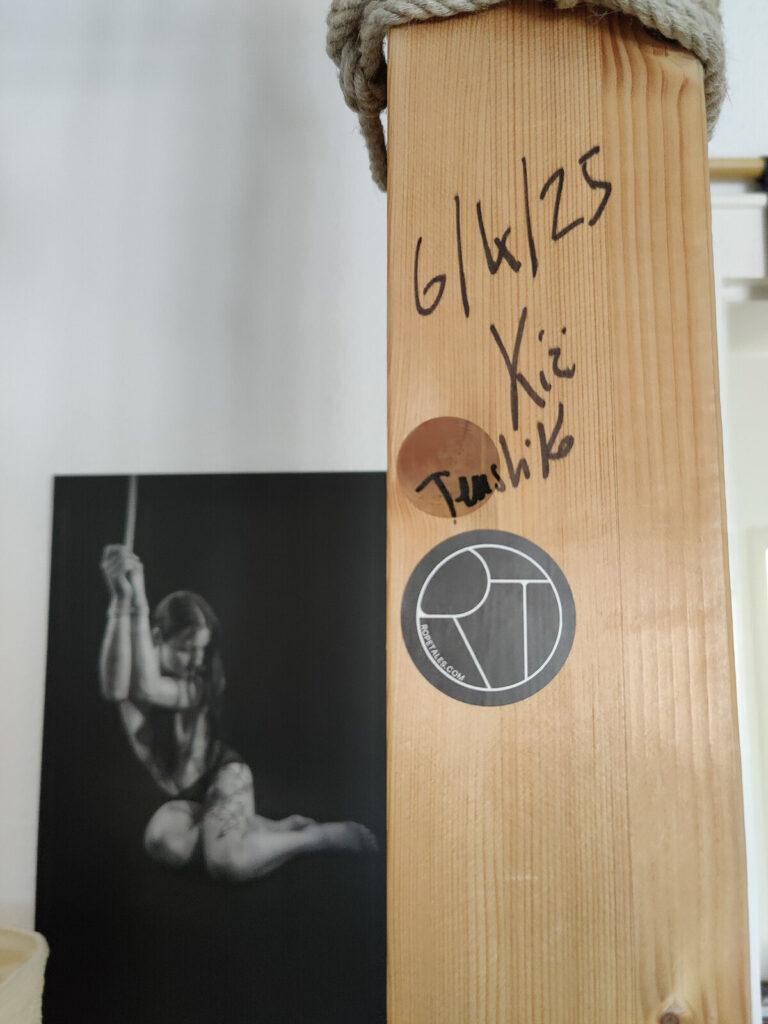 Holzpfosten mit handschriftlicher Widmung und Unterschrift von Kirigami und Tenshiko vom 6. April 2025, im Hintergrund ein Schwarzweißfoto einer Hängefesselung