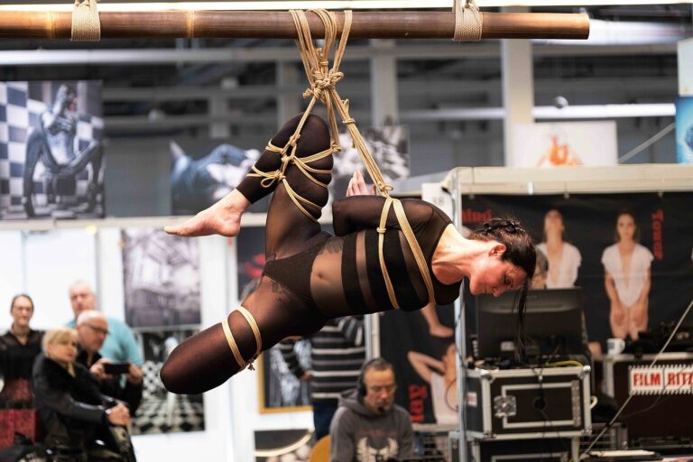 Hängefesselung in Seitenlage im schwarzen Outfit am Bambus, ein Fuß gestreckt, Bondage-Fetish-World Spring 2023