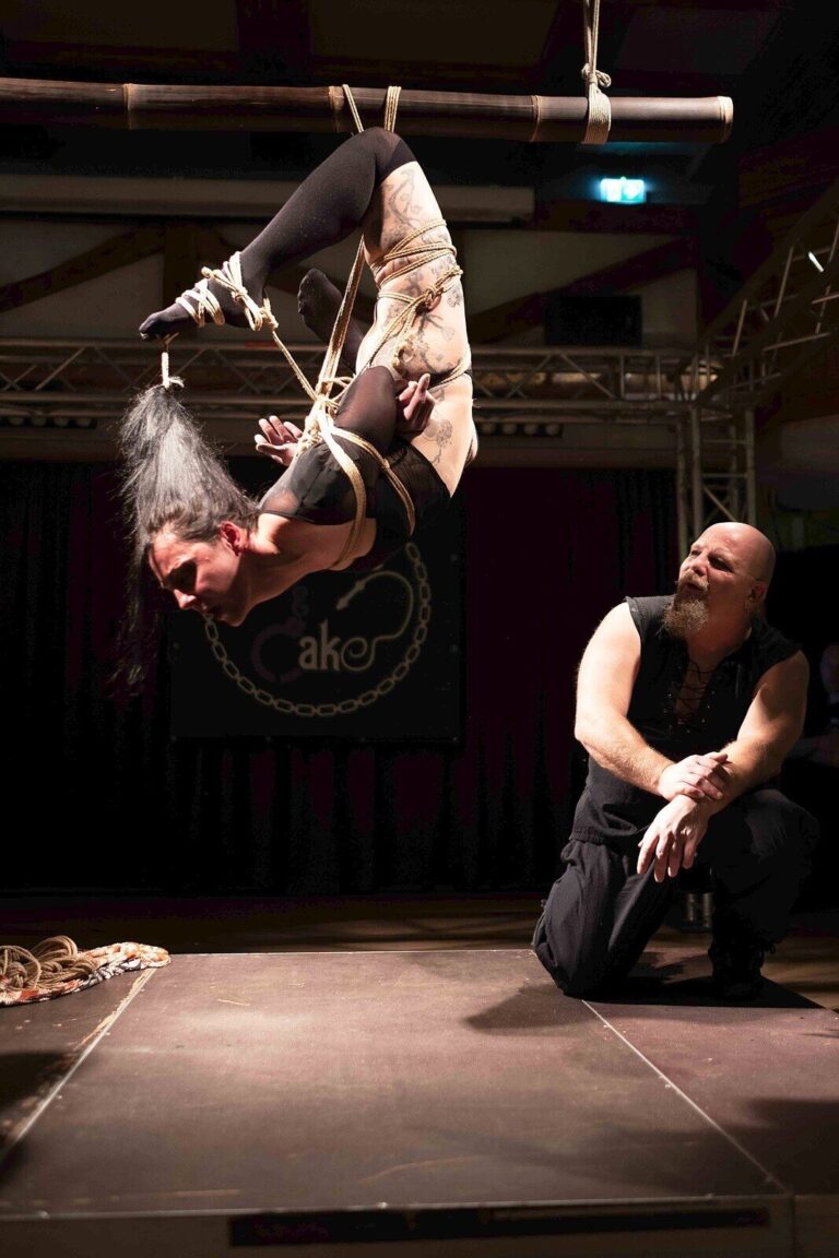 Kopfüber-Hängefesselung im Spotlicht, der Rigger kniet daneben und schaut zu, Fesselnde Nacht