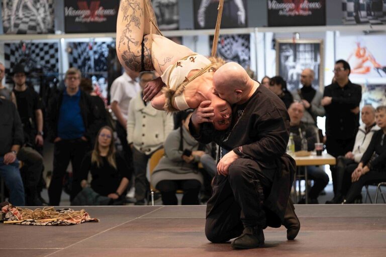 Das Model hängt kopfüber im Fell-Kostüm, der kniende Rigger küsst sie, Publikum im Hintergrund, Bondage-Fetish-World Spring 2023