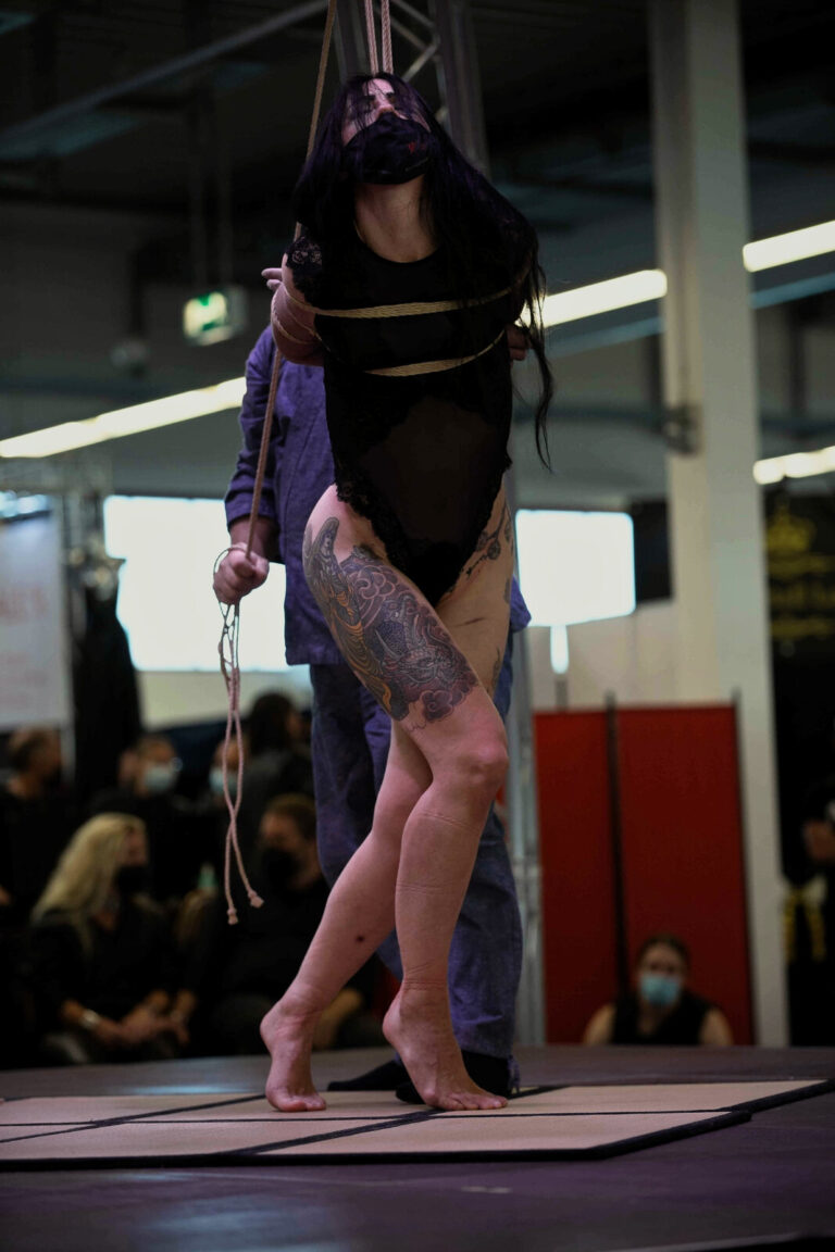 Das Model steht auf Zehenspitzen im Seil, der Rigger hält es von hinten, Bühne der Bondage-Fetish-World 2021