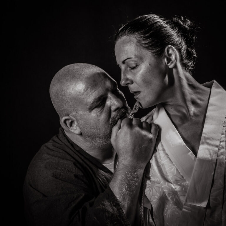 Rigger und Model im weißen Kimono ganz nah beieinander, ein Seil an den Lippen