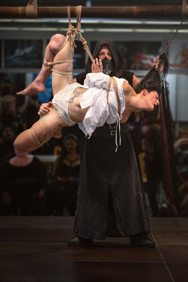 Der Rigger mit schwarzer Kapuze hält das Model im weißen Kleid rücklings in einer Teilhängefesselung am Bambus, Bondage-Fetish-World