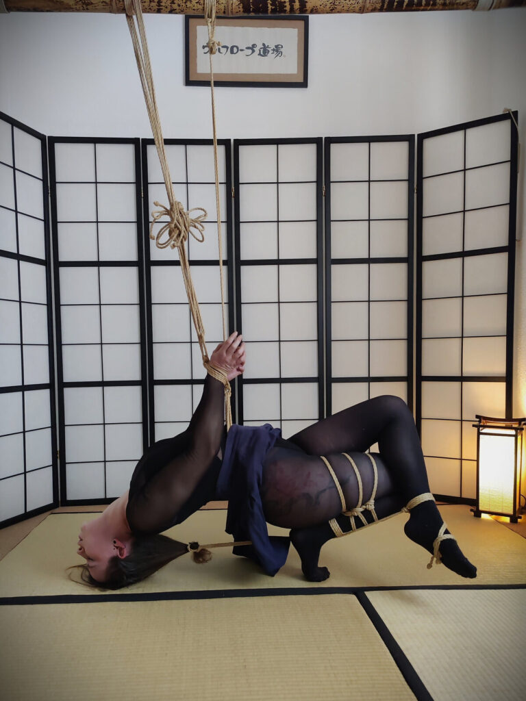 Rücklings auf der Tatami, ein Arm nach oben gebunden, die Beine gefesselt