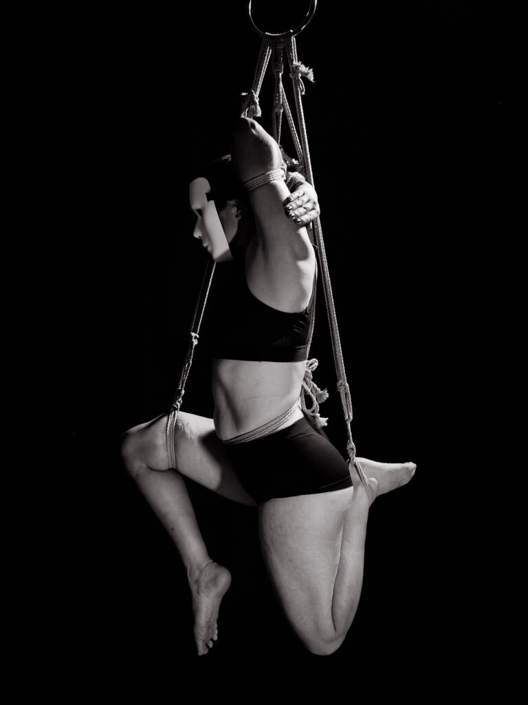 Teilsuspension mit Maske und Sporttop, ein Bein hochgebunden, vor schwarzem Hintergrund